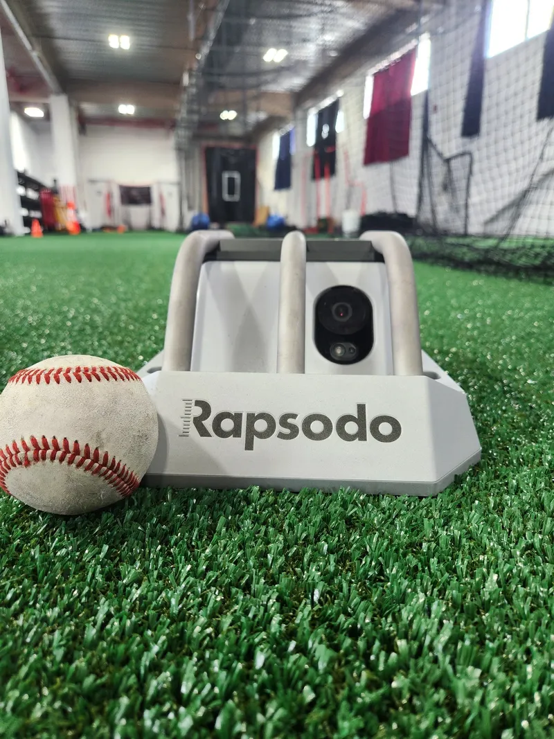 A Rapsodo pitching device next to a baseball on artificial turf.